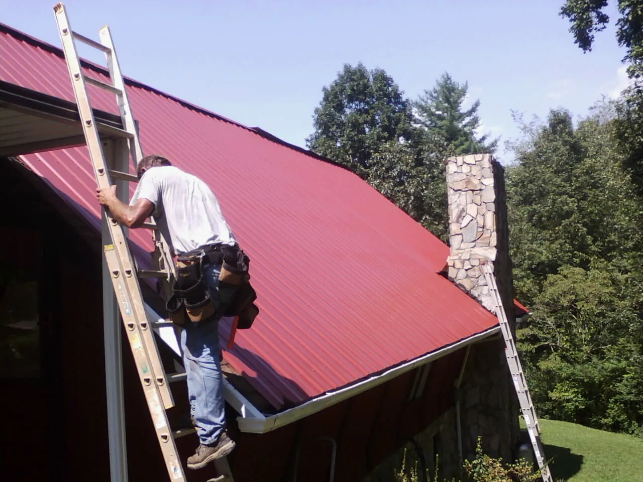 Roofer on a ladder beside a red metal roof and stone chimney — Soffit & Fascia Repair by Roofing Brothers in Dupo